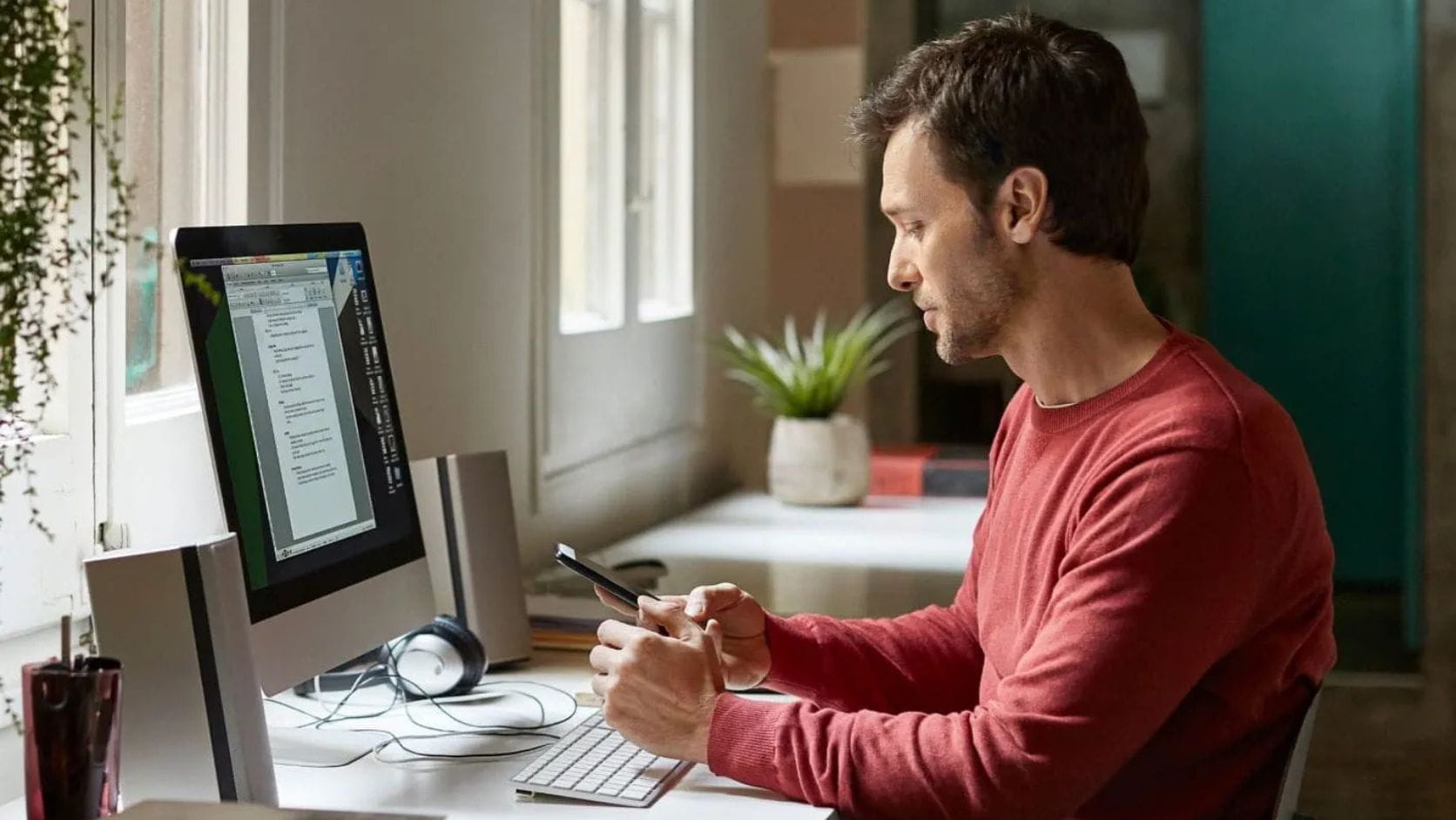Homem trabalha num computador de secretária em casa enquanto consulta o telemóvel, analisando opções num simulador para saber quanto pode pedir de crédito habitação e planear o financiamento.