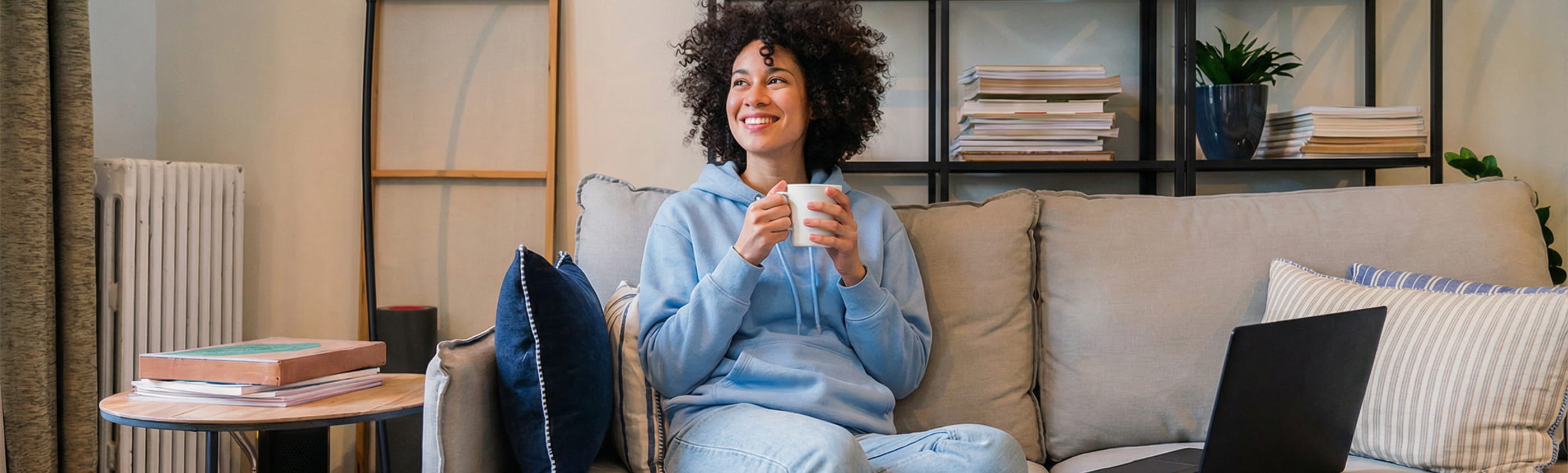 Mulher sentada confortavelmente no sofá em casa com uma chávena e um portátil ao lado, cenário associado à tranquilidade após terminar o crédito habitação e ao processo de distrate da hipoteca.
