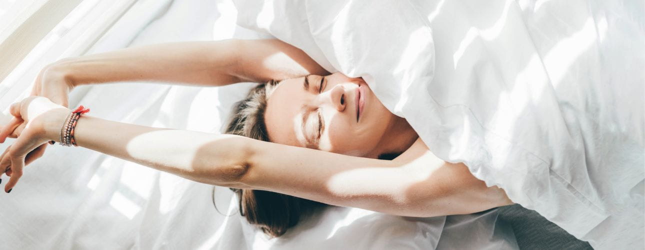 Mulher deitada numa cama com lençóis brancos junto a uma janela com luz natural, cenário associado ao conforto da habitação e à tranquilidade financeira proporcionada por uma solução de crédito habitação.