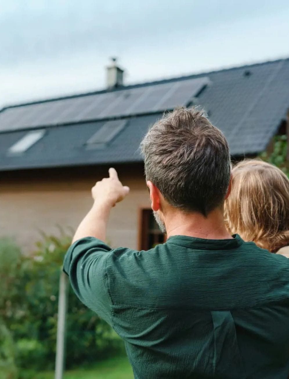 Um pai com a filha ao colo a observar uma moradia equipada com painéis solares no telhado, cenário associado à eficiência energética da habitação, à sustentabilidade e às soluções de crédito habitação para construção de casas energeticamente eficientes.